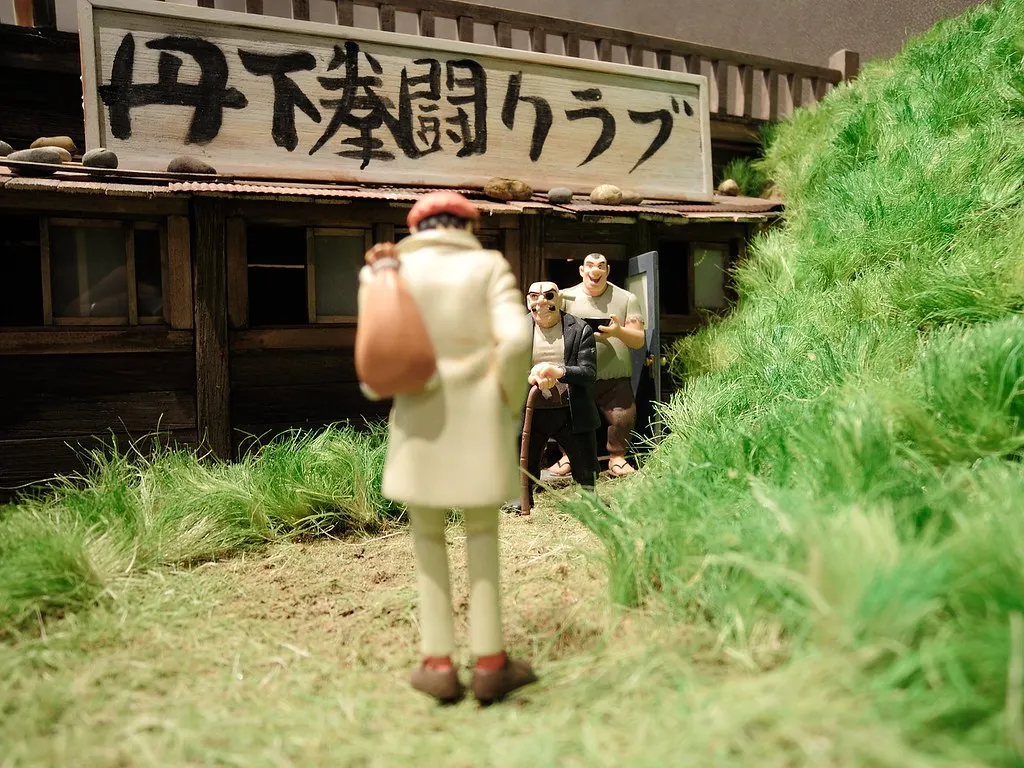Diorama de figurines devant une baraque en bois portant l'enseigne « 丹下拳闘クラブ ». Vu de dos, un jeune homme en manteau clair, sac sur l'épaule, fait face à un vieil homme borgne appuyé sur une canne et à un homme corpulent tenant un bol.