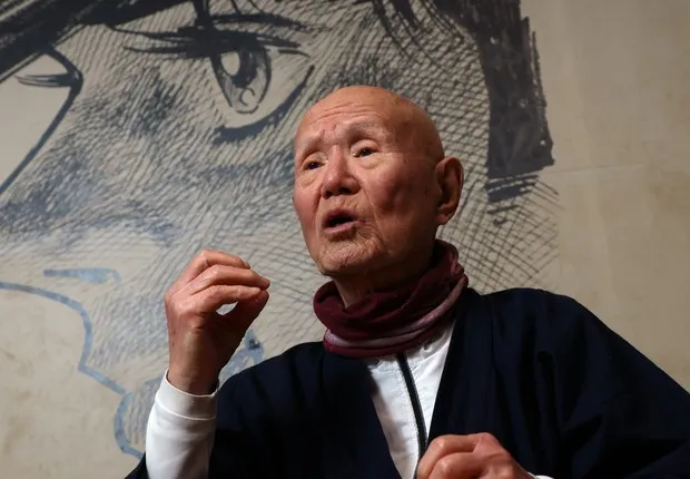 Portrait de Tetsuya Chiba en train de parler, la main levée, vêtu d'un haut sombre et d'une écharpe bordeaux, devant un grand dessin en noir et blanc d'un visage de manga dont on voit surtout un œil