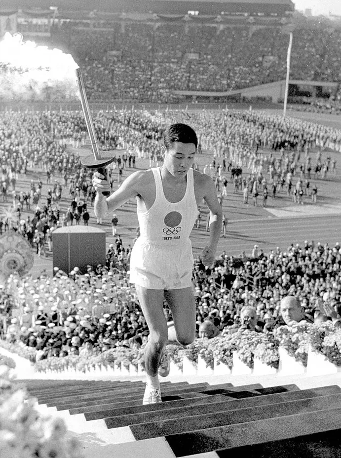 Photographie en noir et blanc d'un jeune coureur en maillot blanc frappé du disque solaire et des anneaux olympiques, inscription « TOKYO 1964 », gravissant des marches torche enflammée à la main, devant les tribunes bondées d'un stade.