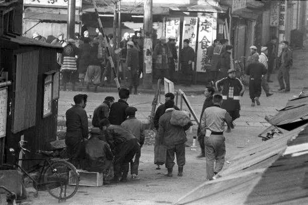 Photographie en noir et blanc d'un carrefour de Sanya en hiver : des hommes en vestes et casquettes, certains chargés de sacs, attendent ou passent entre des baraques aux toits de tôle, devant des enseignes de petits restaurants.