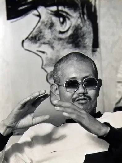 Photographie en noir et blanc d'Ikki Kajiwara, crâne rasé, moustache et lunettes teintées, qui parle en gesticulant des deux mains, assis devant un grand dessin de profil d'un personnage de manga.