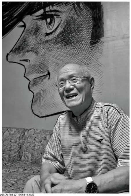 Photographie en noir et blanc de Tetsuya Chiba, souriant, lunettes et polo rayé, assis devant un immense dessin à l'encre du profil de Joe Yabuki.