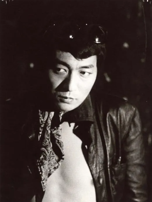 Portrait en noir et blanc de Shūji Terayama sur fond sombre, lunettes de soleil relevées dans les cheveux, blouson de cuir ouvert sur un foulard à motifs cachemire, le regard tourné de côté