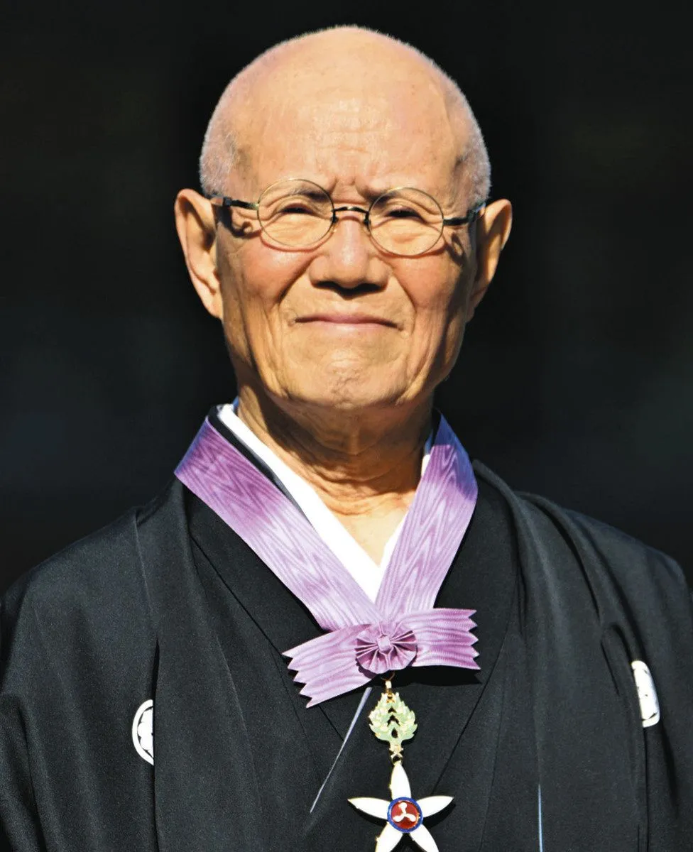 Portrait d'un homme âgé au crâne rasé et aux lunettes rondes, souriant, en kimono noir à blason, portant au cou l'insigne de l'Ordre de la Culture suspendu à un ruban violet.