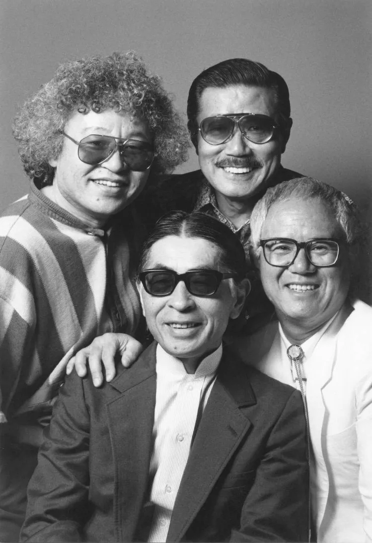 Portrait de groupe en noir et blanc de quatre mangakas souriants : Shōtarō Ishinomori, cheveux bouclés et lunettes teintées, Takao Saitō, moustache et lunettes d'aviateur, Fujiko Fujio Ⓐ, lunettes noires et veste sombre, et Tetsuya Chiba, lunettes et veste claire.