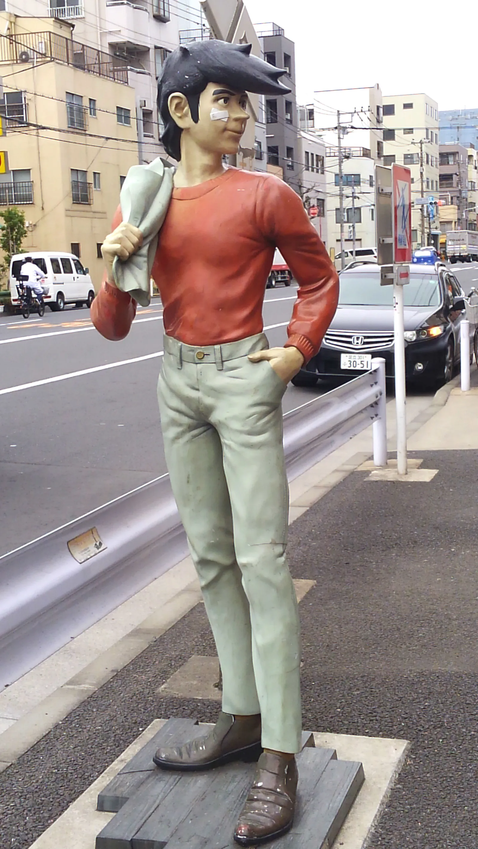 Statue peinte de Joe Yabuki, cheveux noirs en bataille et pansement sur la joue, pull rouge et pantalon clair, une veste sur l'épaule et une main dans la poche, sur un trottoir au bord d'une avenue de Sanya.