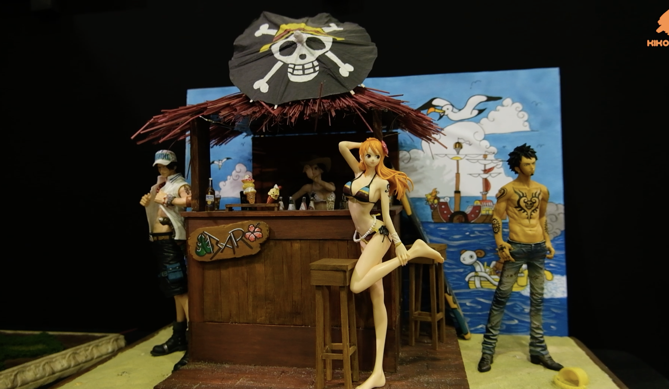 Diorama One Piece&nbsp;: Nami et Trafalgar Law devant un bar de plage surmonté d’un drapeau pirate