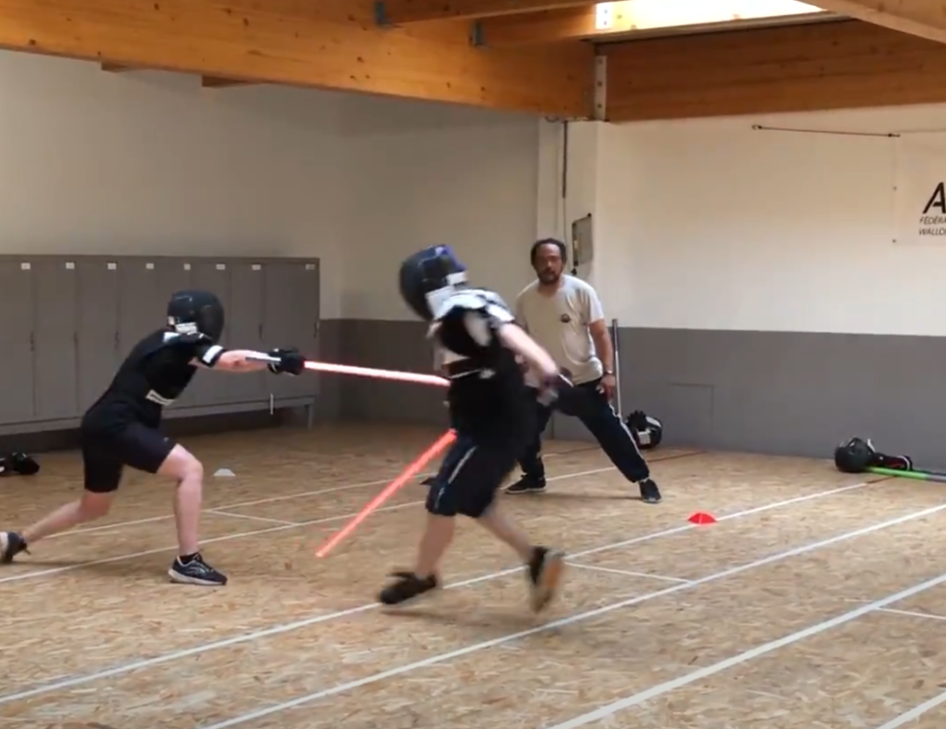 Deux pratiquants casqués croisent leurs sabres laser rouges sous le regard d’un arbitre