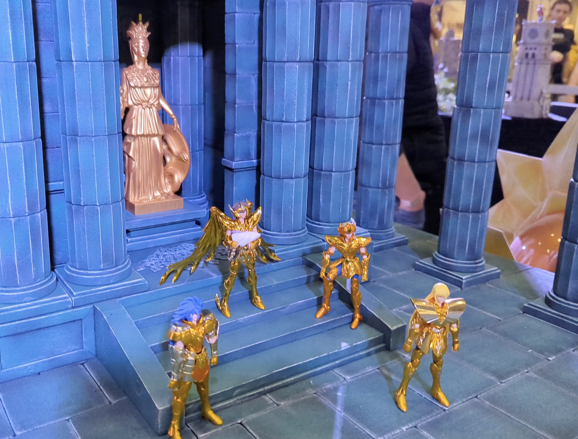 Figurines de chevaliers d’or sur les marches d’un temple bleu, sous une statue dorée d’Athéna