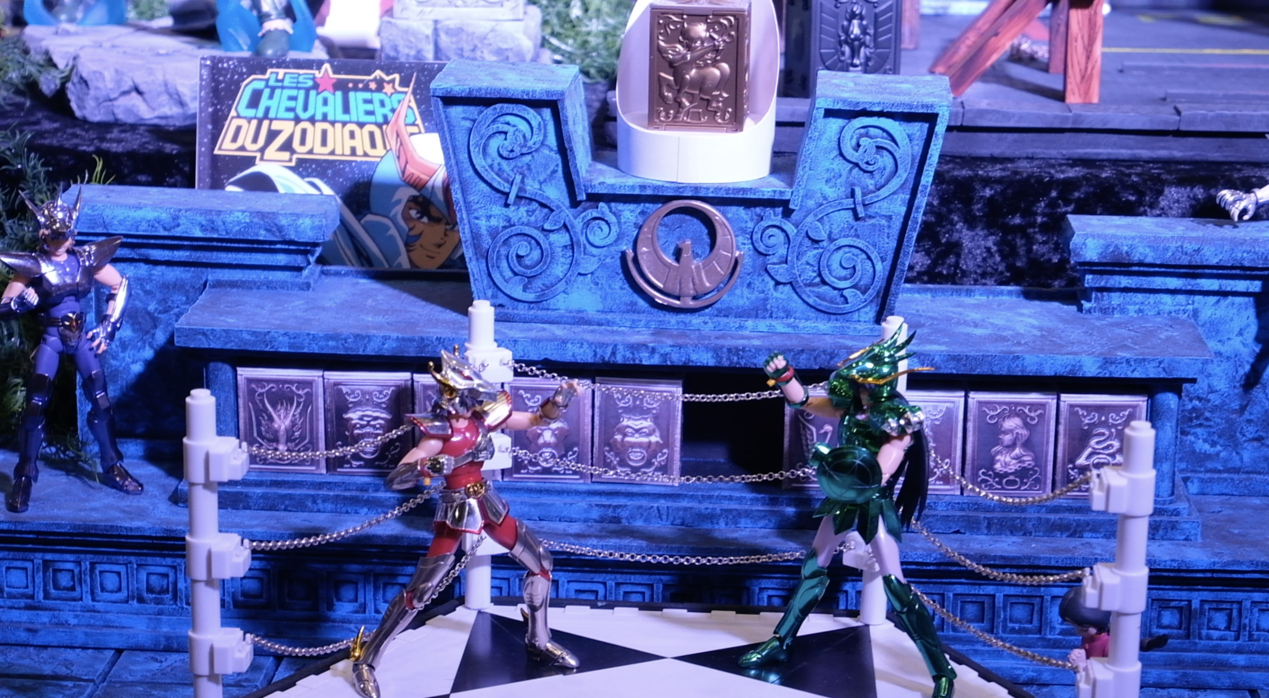 Figurines de Seiya et Shiryu face à face sur un ring à damier, devant les boîtes d’armures