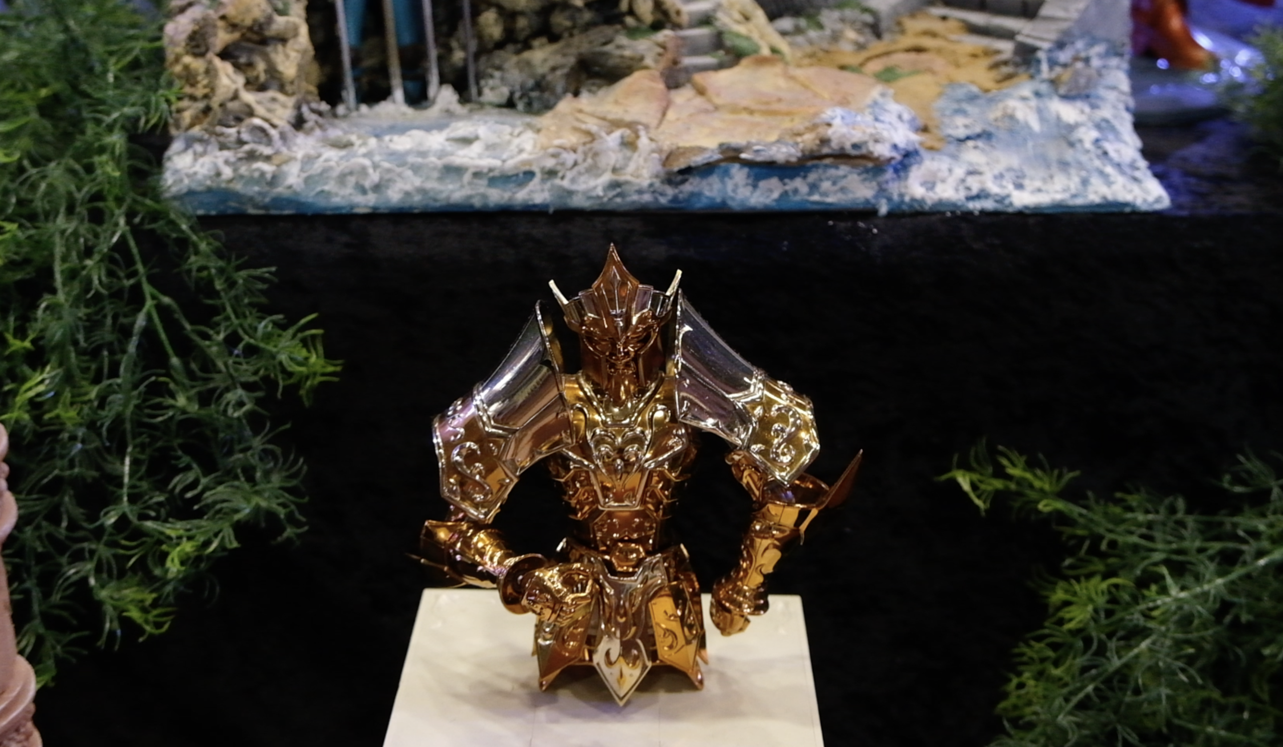 Gros plan sur une figurine en armure dorée posée sur un socle blanc, devant un décor rocheux