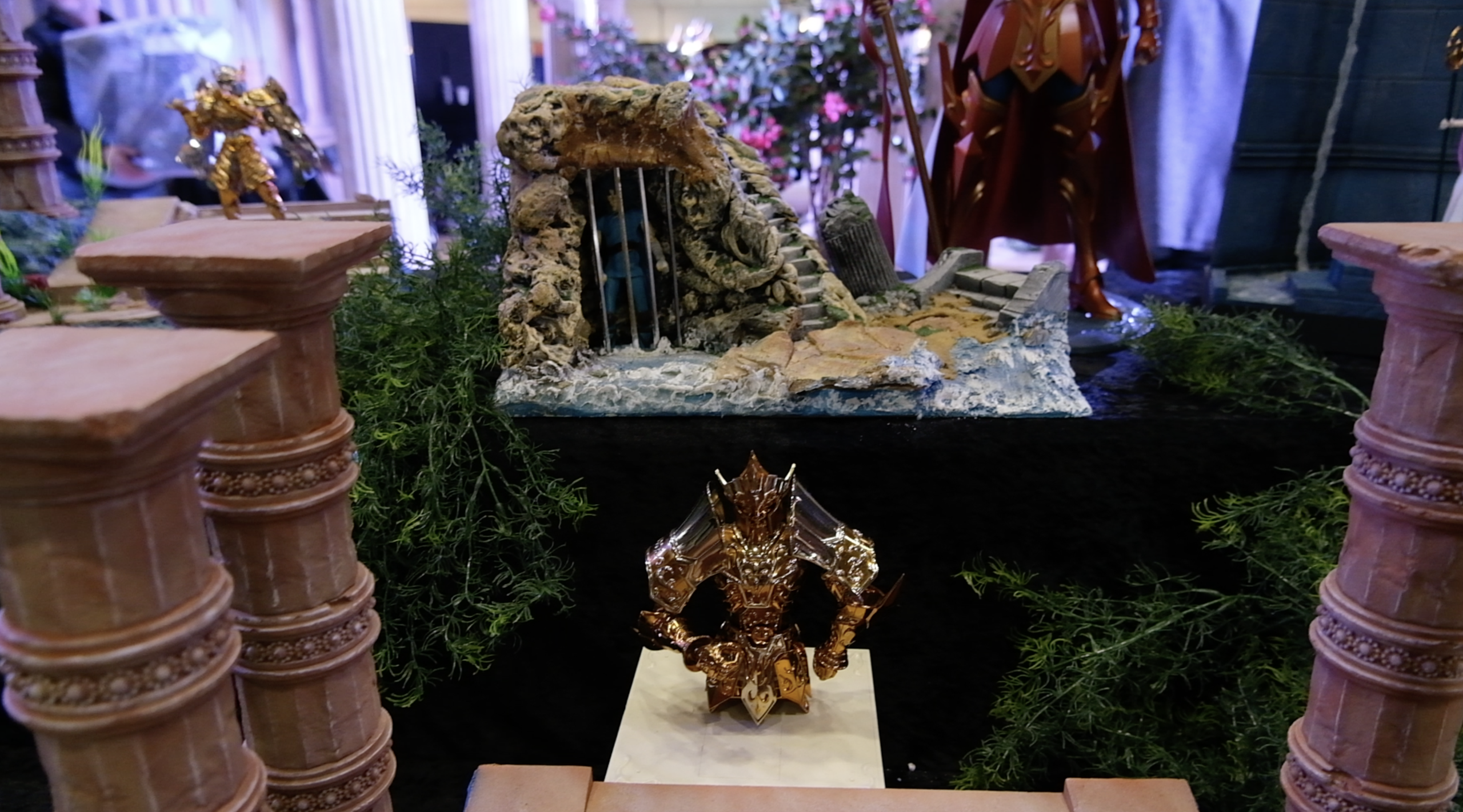 Figurine en armure dorée sur un socle, devant une prison rocheuse miniature et des colonnes