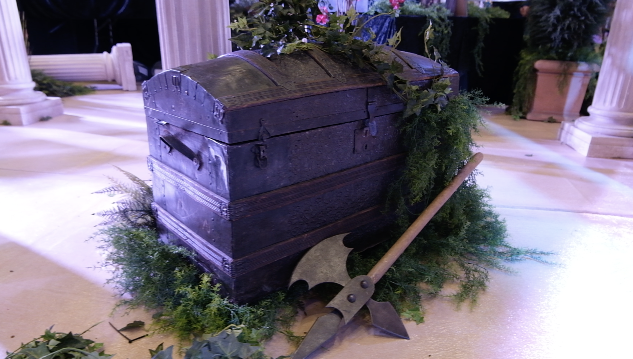 Vieille malle en bois et hallebarde posées dans la verdure, au pied des colonnes du décor