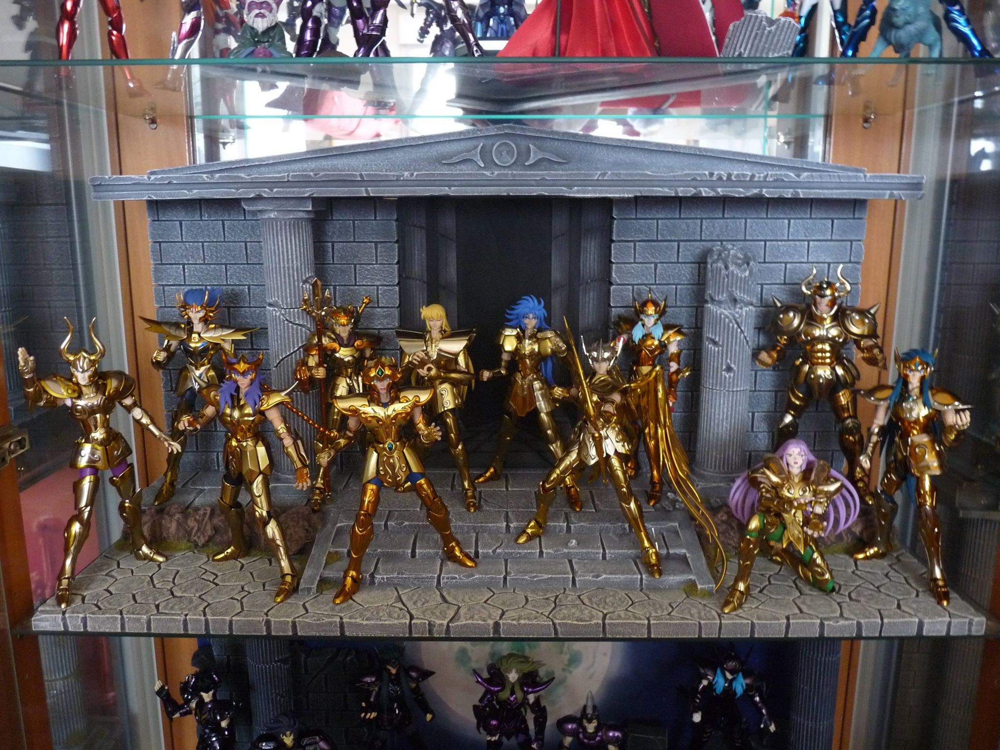 Figurines des chevaliers d’or de Saint Seiya devant un temple grec, dans une vitrine