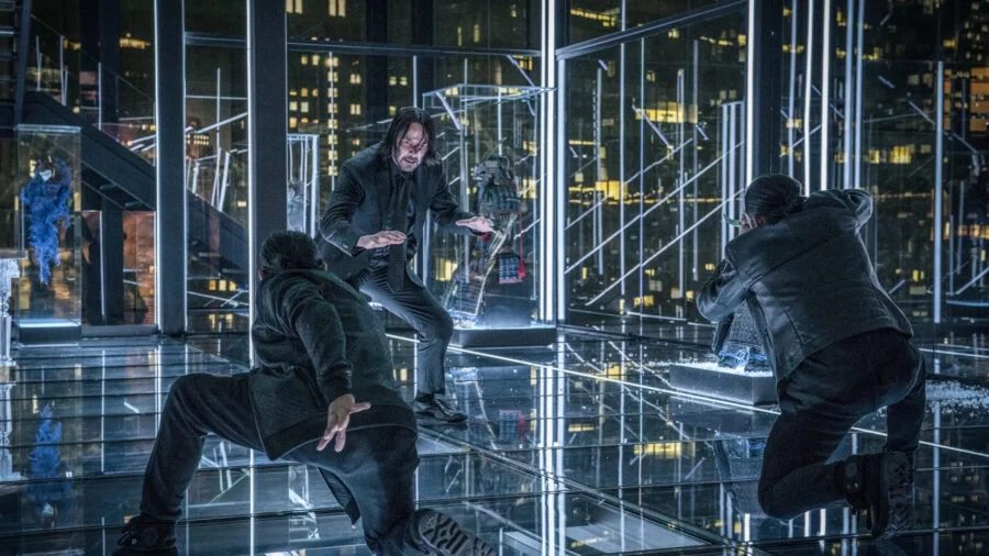 John Wick face à deux assaillants dans une salle aux murs de verre surplombant la ville