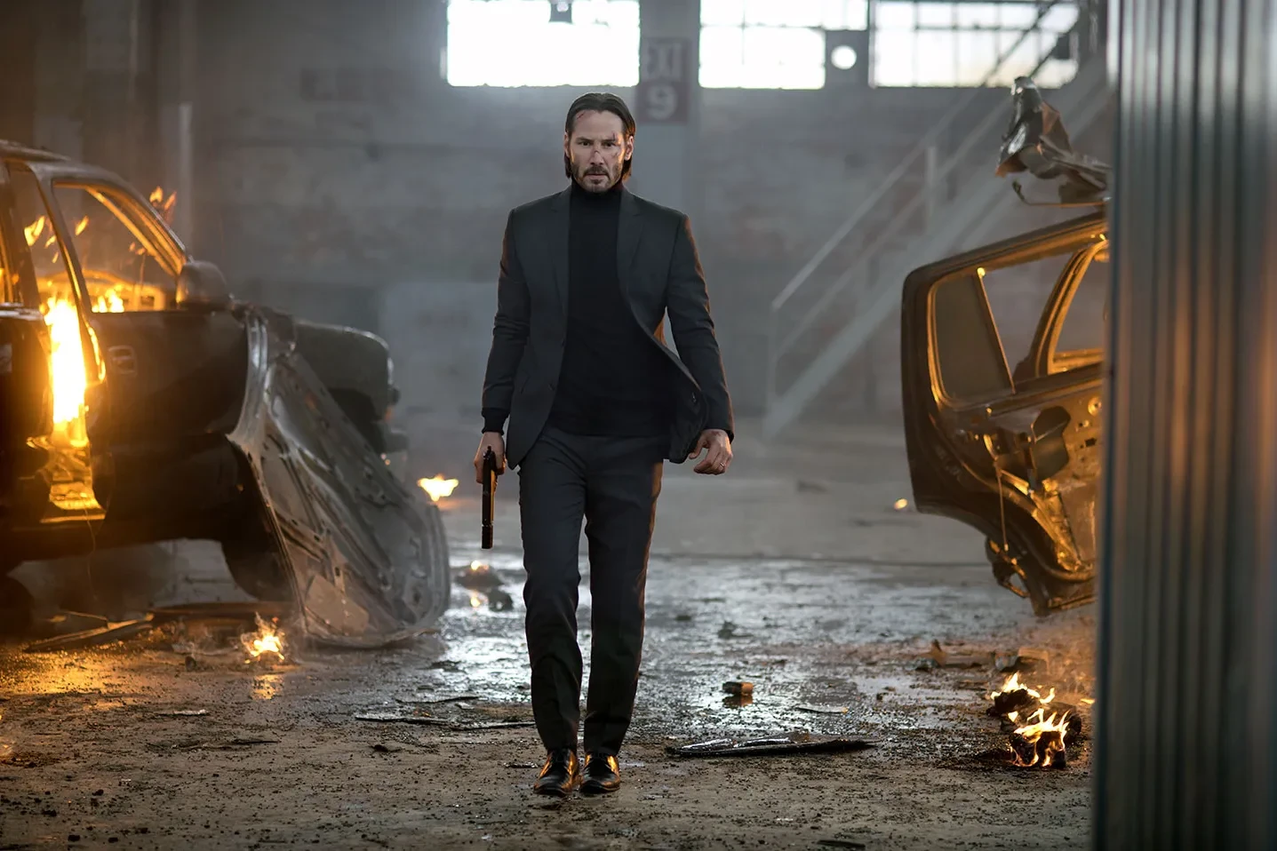 John Wick, pistolet à la main, avance entre des carcasses de voitures en feu dans un entrepôt