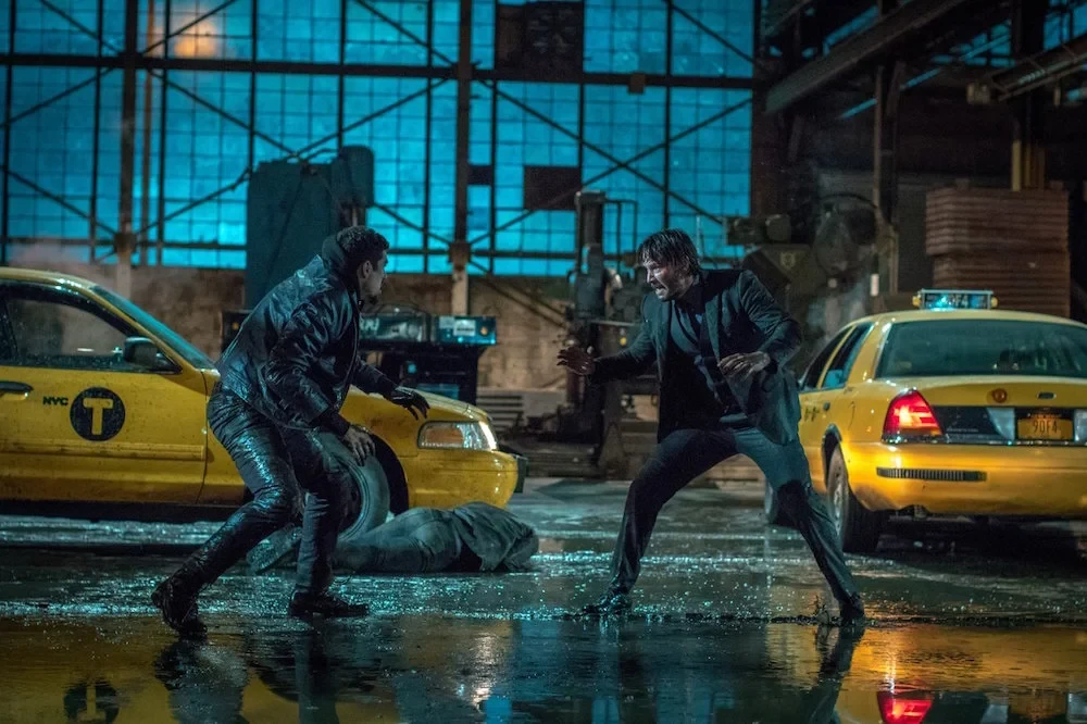 John Wick affronte un homme en cuir entre deux taxis jaunes, dans un entrepôt détrempé