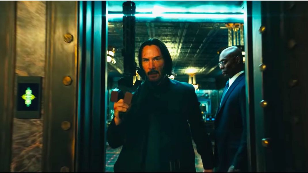 John Wick, fusil à l’épaule, sort d’une salle forte, suivi de Charon en costume