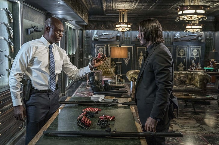Lance Reddick en Charon tend une cartouche à John Wick devant une table couverte d’armes