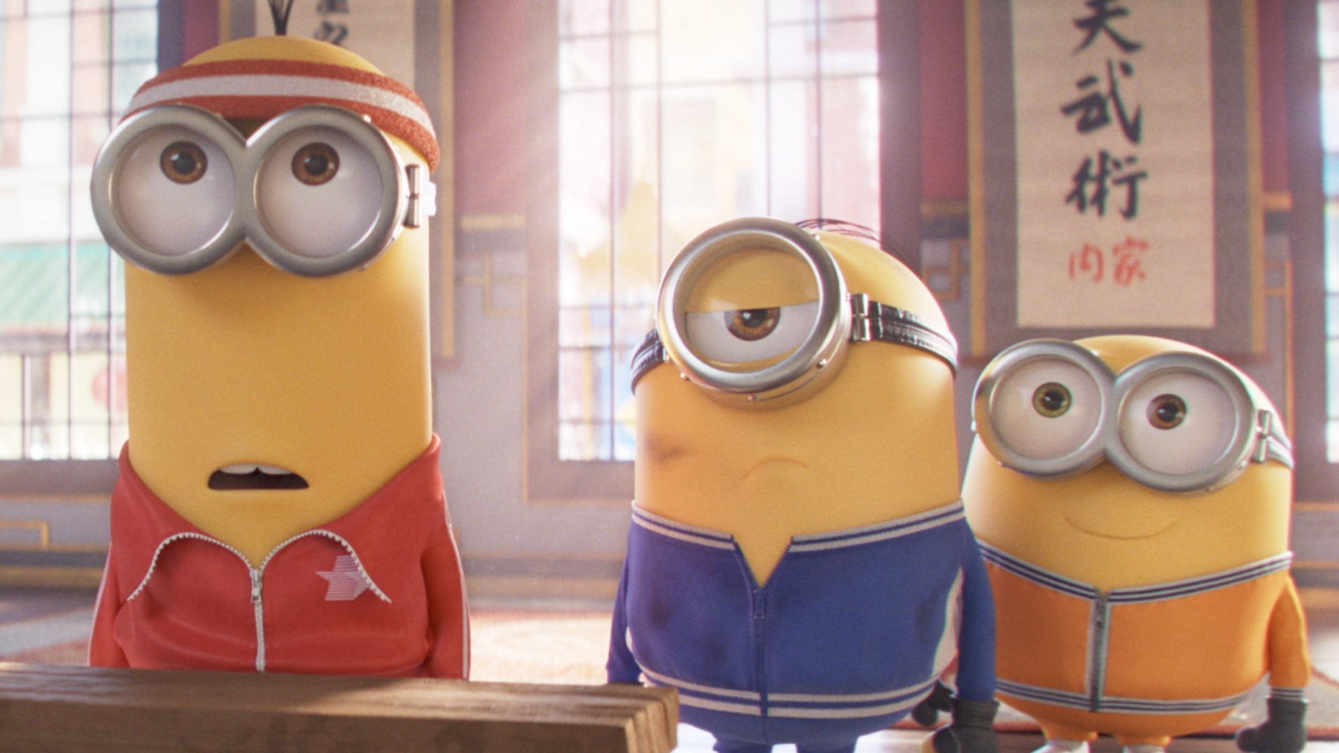 Trois Minions en survêtements rouge, bleu et orange dans un dojo
