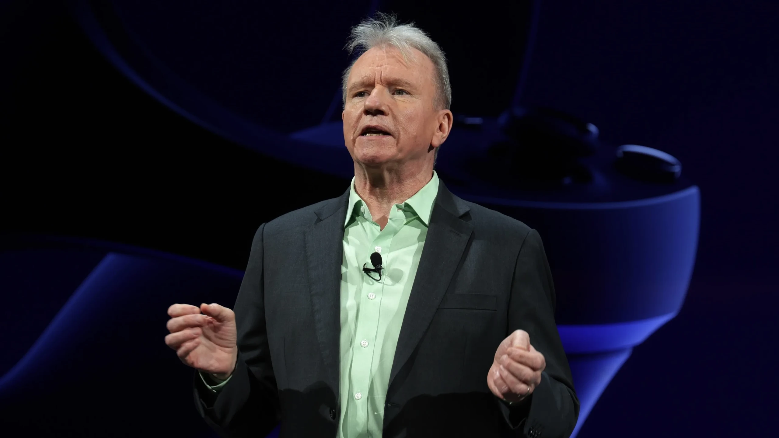 Jim Ryan, en veste sombre et chemise vert clair, s’exprime sur scène devant un fond bleu PlayStation