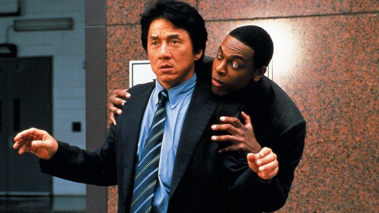 Chris Tucker se cache derrière Jackie Chan, l’air inquiet, contre un mur de granit