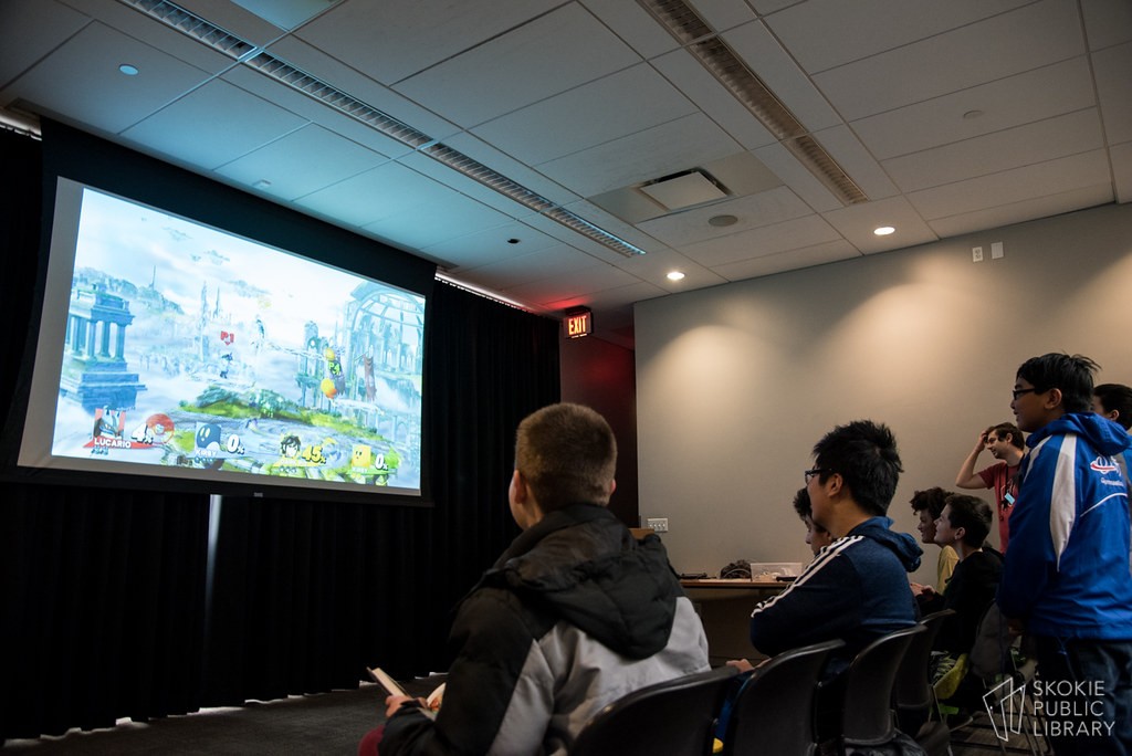 Joueurs assis devant un écran géant diffusant un match de Super Smash Bros. lors d’un tournoi en salle