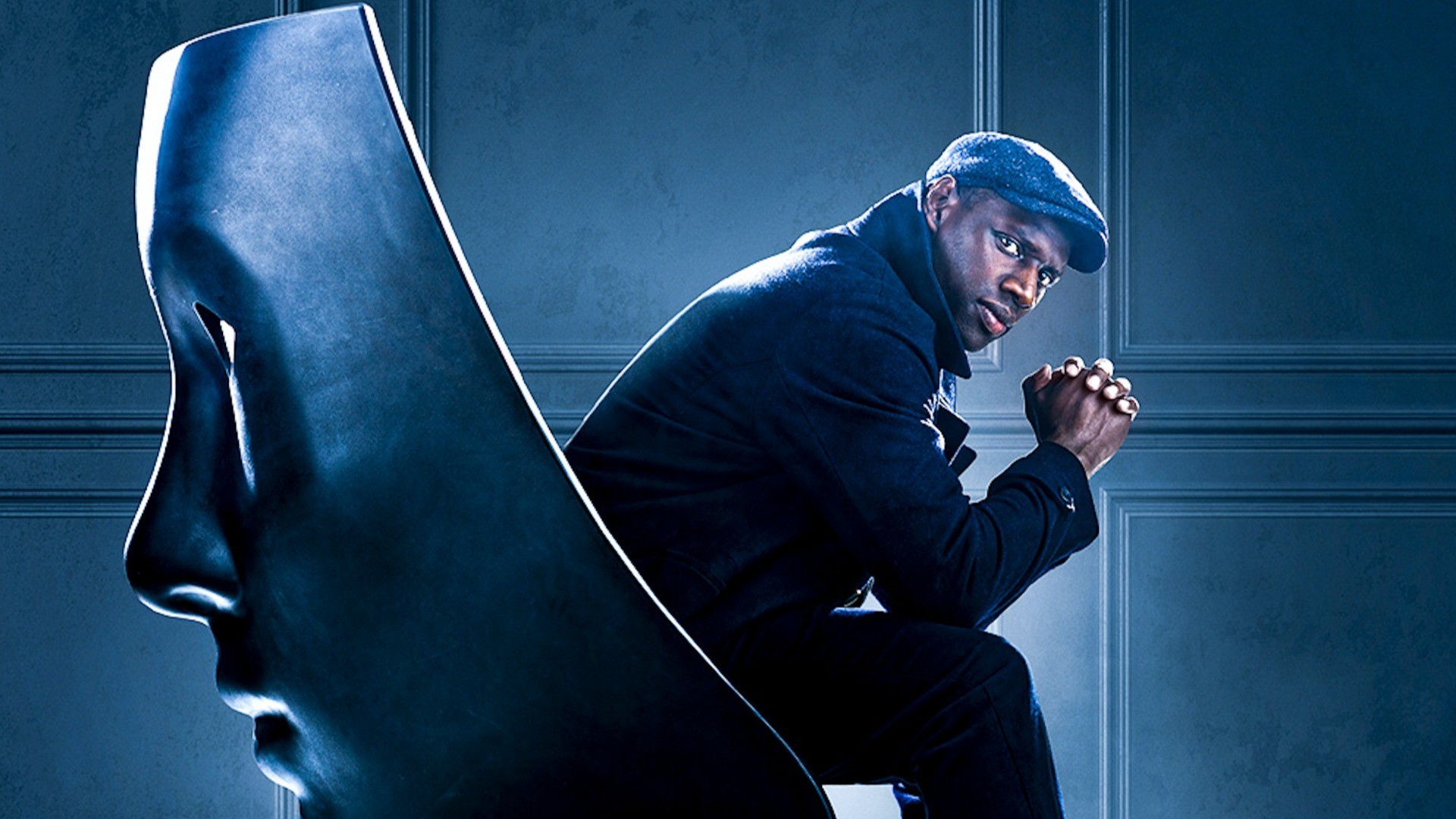 Omar Sy en Assane Diop, casquette sur la tête, assis à côté d’un grand masque