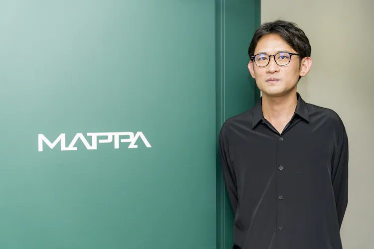 Homme à lunettes en chemise noire, posant à côté du logo MAPPA sur un mur vert