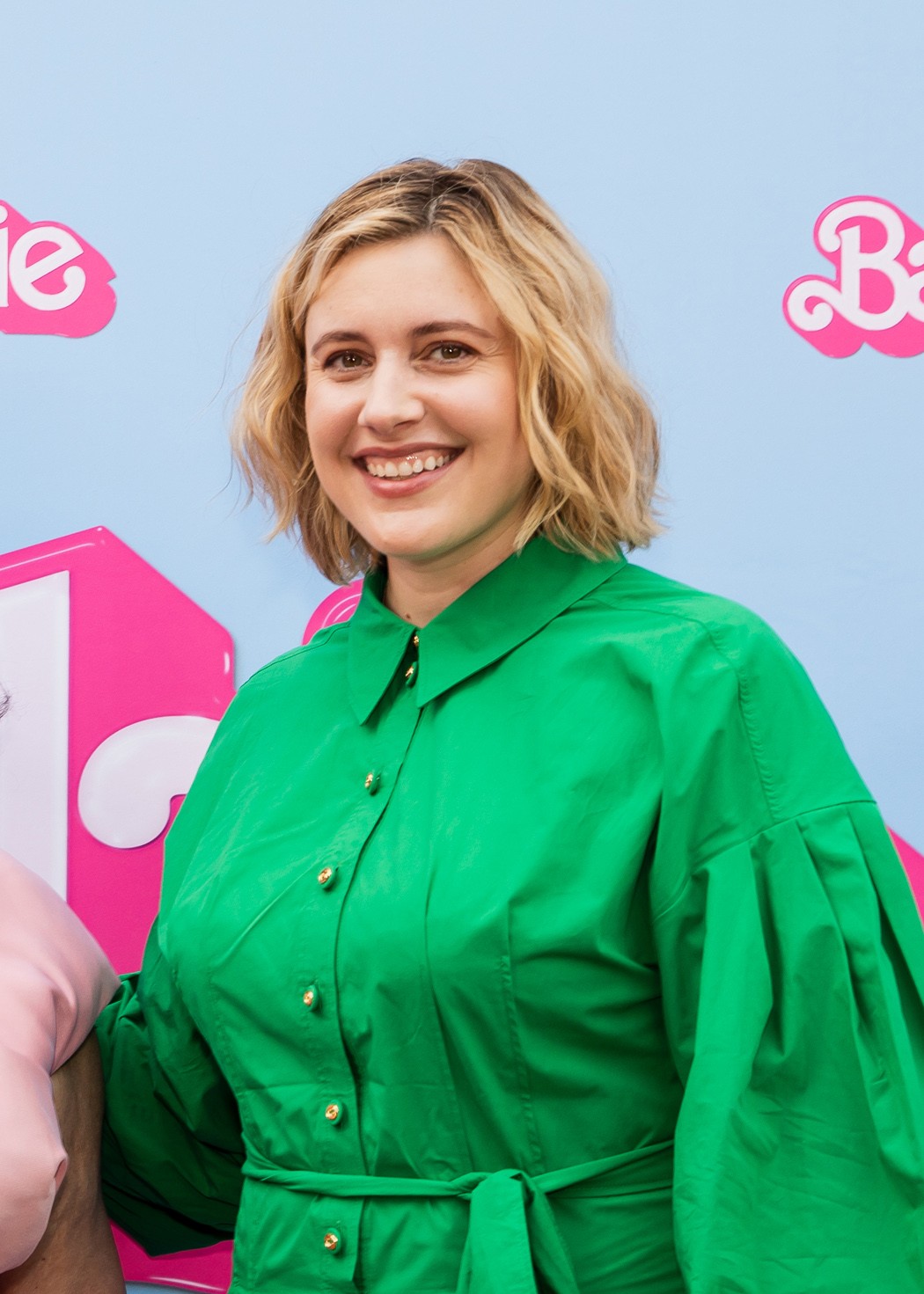 Greta Gerwig, souriante en chemise verte, devant un mur aux logos Barbie