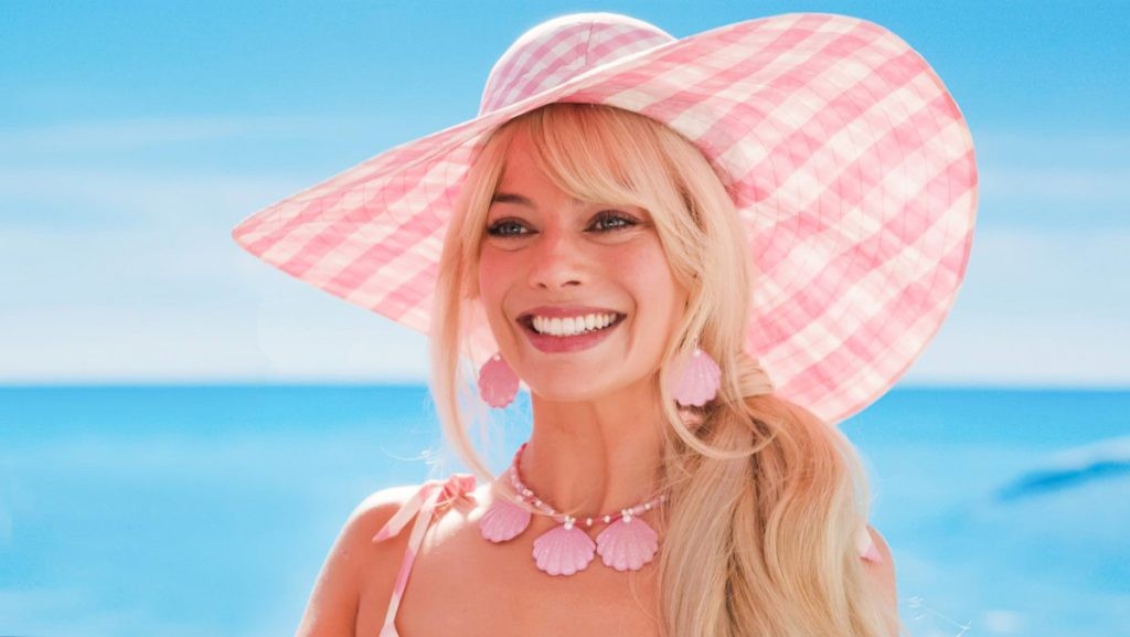 Barbie, jouée par Margot Robbie, sourit sous un grand chapeau rose à la plage