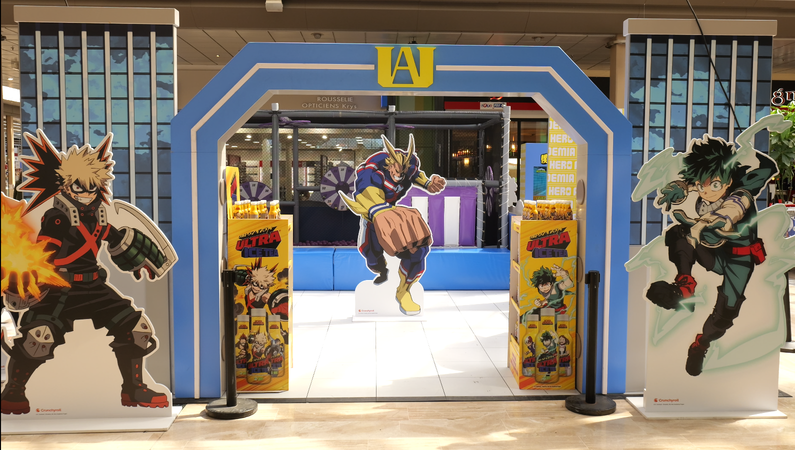 Portique aux couleurs du lycée Yuei entre des silhouettes de Bakugo et Deku dans un centre commercial